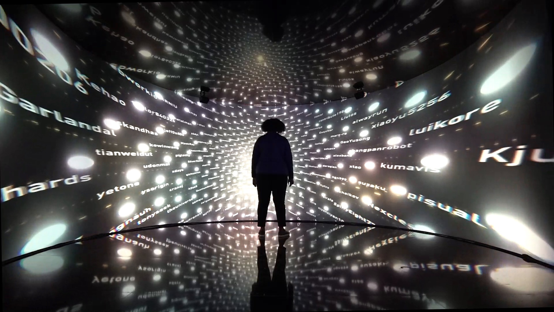 A person stands silhouetted inside Cerebro, a curved tunnel of screens at GitHub Satellite 2019 in Berlin. Hundreds of glowing dots and GitHub usernames surround them on every side and reflect off the floor.